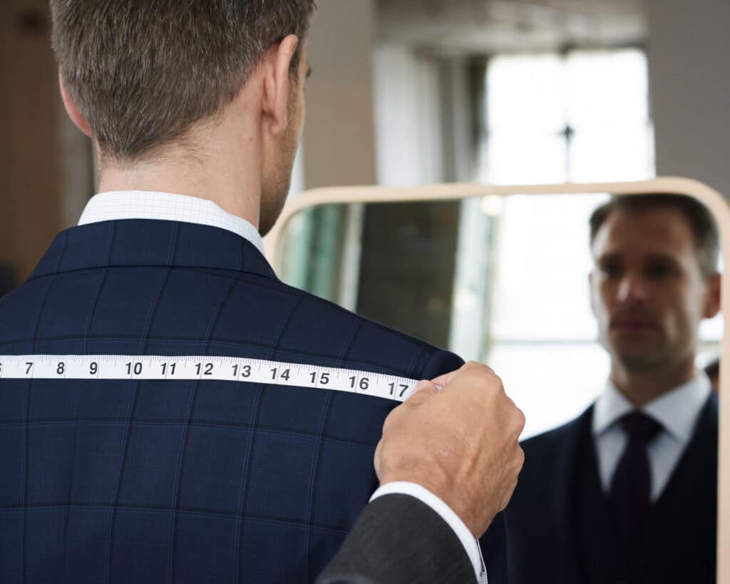 Custom tailor measuring to point to point shoulder width on a client in front of the mirror