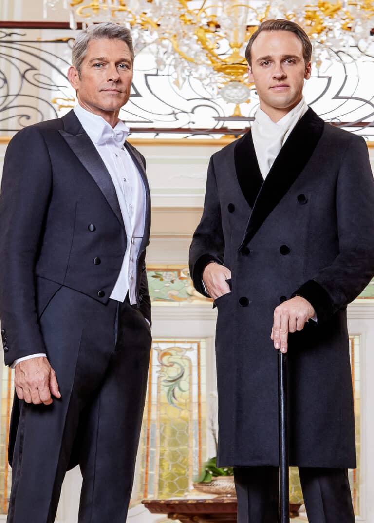 Two guests standing at base of majestic staircase. One wearing bespoke tuxedo tails and one wearing double breasted bespoke overcoat with white scarf.