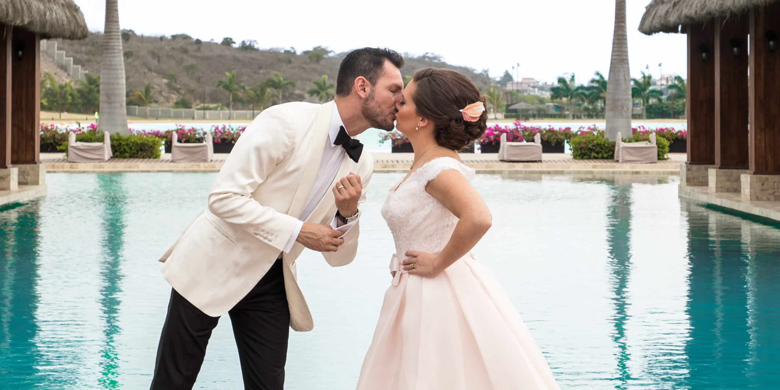 Groom kissing bride in front of pool. Groom wearing Loro Piana white bespoke dinner jacket with shawl lapel. Bride in white sleeveless dress.