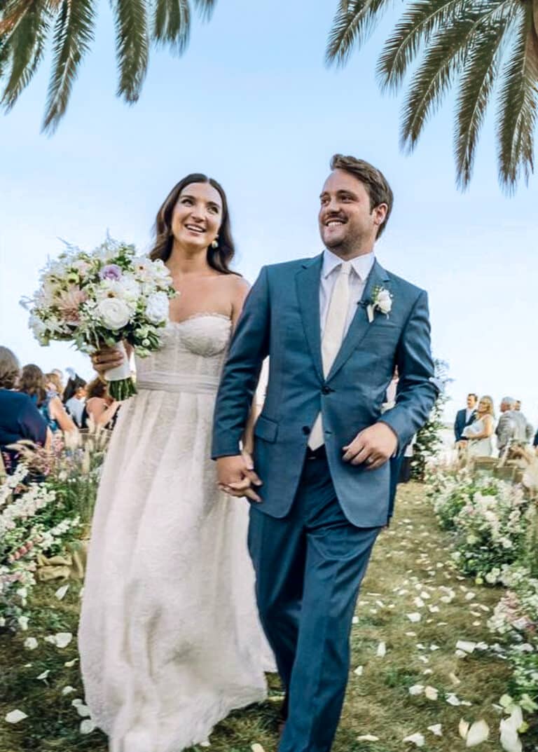 Bride and groom on beach holding hands. Groom in postman blue Zegna custom suit, bride in low cut wedding dress.