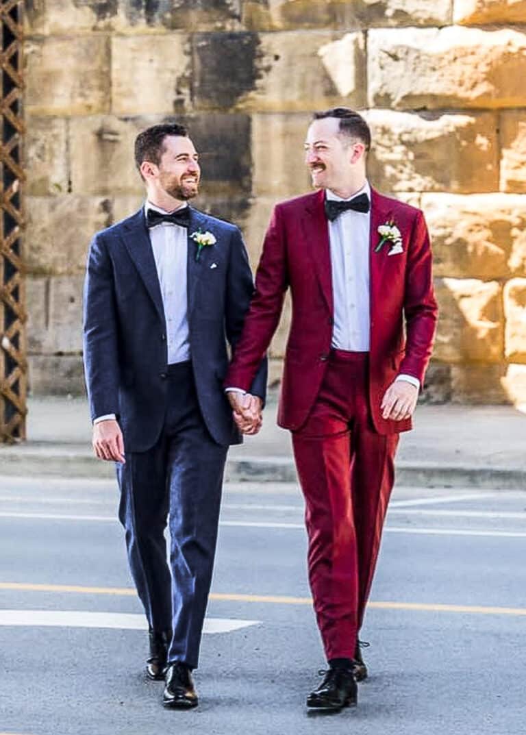 Groom and groom holding hands. One wearing solid dark blue custom wedding suit with white shirt and wide blue bowtie. Other wearing red solid bespoke wedding suit with white shirt and oversized black bowtie.