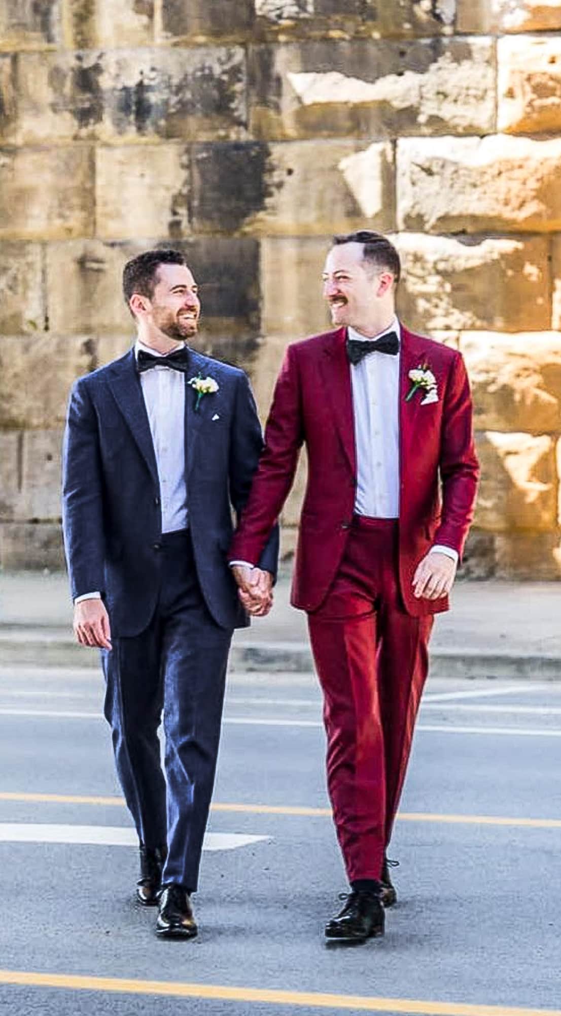 Groom and groom holding hands. One wearing solid dark blue custom wedding suit with white shirt and wide blue bowtie. Other wearing red solid bespoke wedding suit with white shirt and oversized black bowtie.