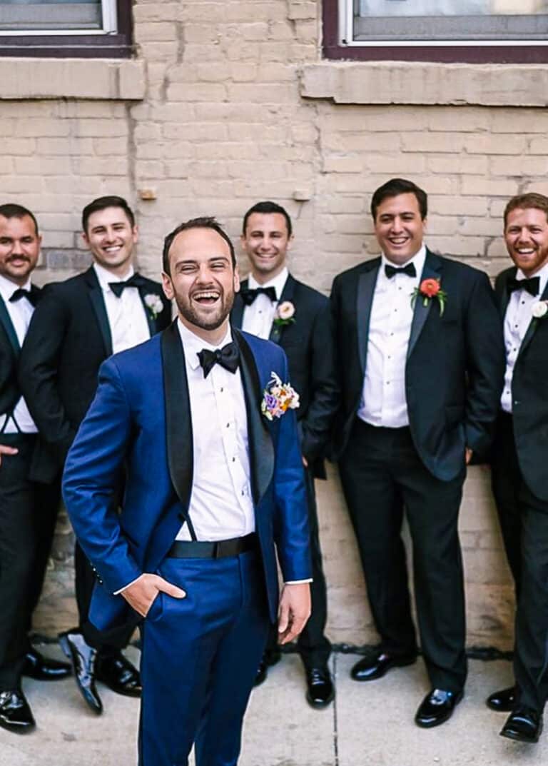 Five groomsmen standing behind groom in black custom tailored tuxedos. Groom wearing blue solid sharkskin tuxedo with black shawl lapel.