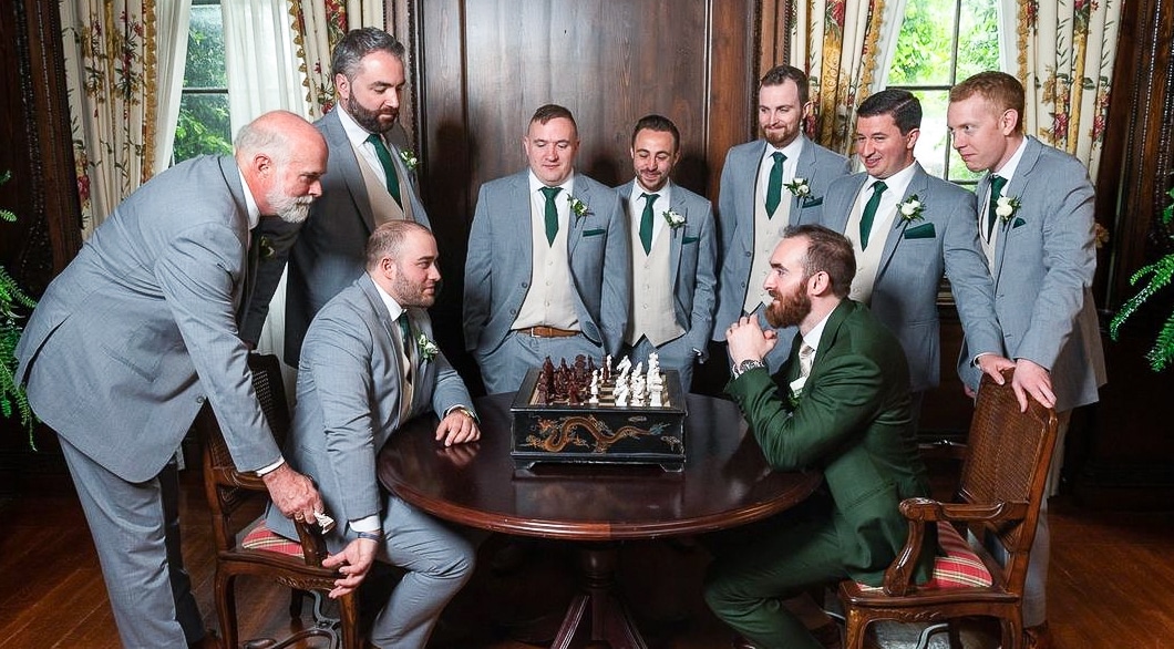 Nine groomsmen wearing very light blue custom wedding suits with white vests, watching 2 groomsmen playing chess.