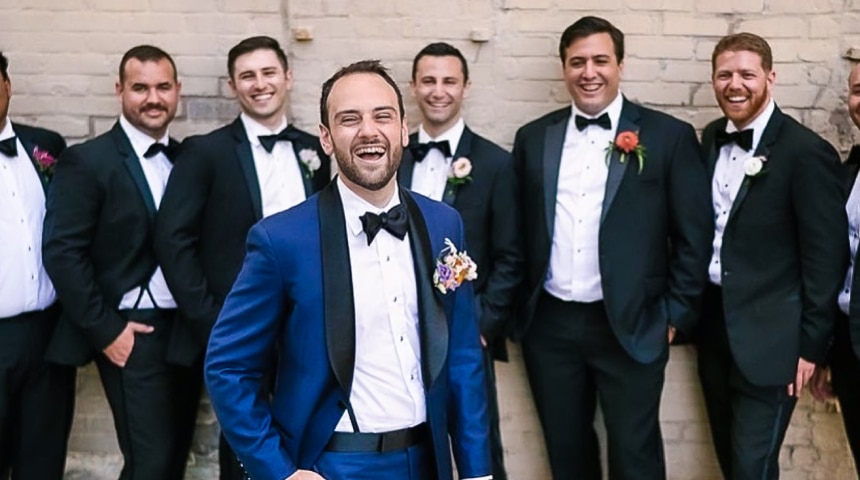 Five groomsmen standing behind groom in black custom tailored tuxedos. Groom wearing blue solid sharkskin tuxedo with black shawl lapel.