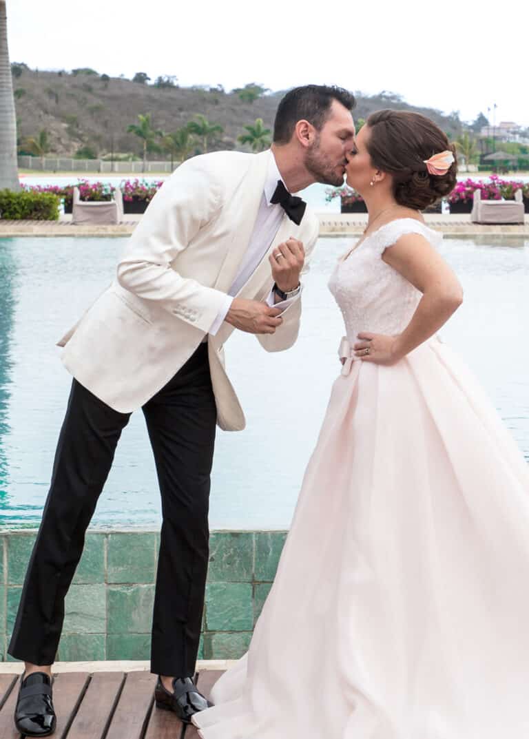 Groom kissing bride in front of pool. Groom wearing Loro Piana white bespoke dinner jacket with shawl lapel. Bride in white sleeveless dress.