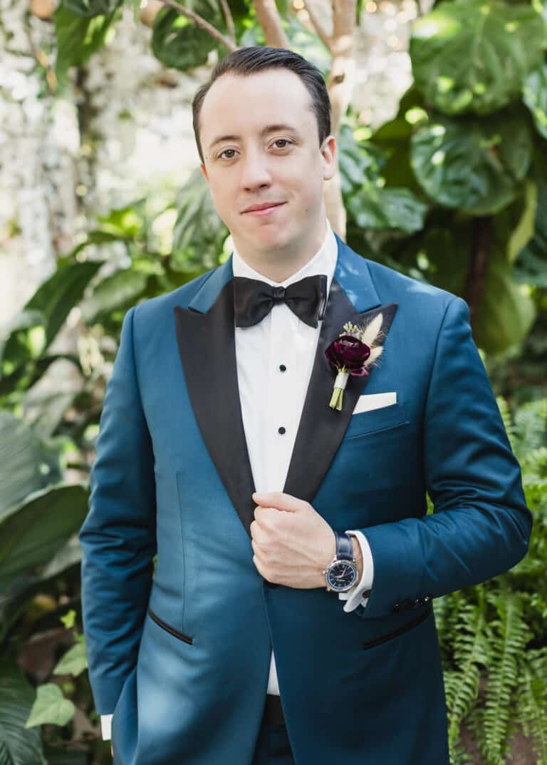 Groom outdoors wearing blue wedding custom tailored tuxedo with black peak lapel. Fabric by Scabal.