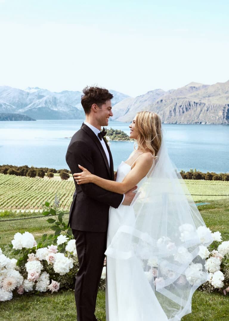 Bride and groom looking at each other with lake and mountains in background. Groom in Vitale Barberis black custom wedding tuxedo, white shirt and black bowtie. Bride in sleeveless wedding dress.