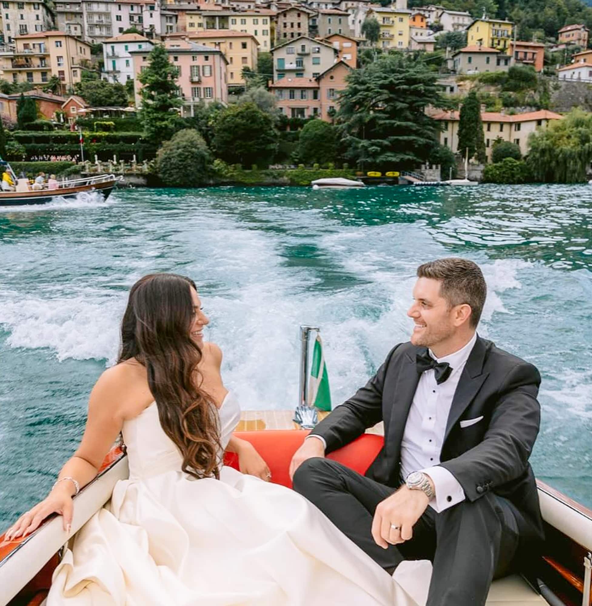 Bride and groom in boat sailing away with Italian beach houses in background. Groom wearing black peak lapel bespoke wedding tuxedo with black satin, white shirt and black bowtie. Bride in sleeveless wedding dress.