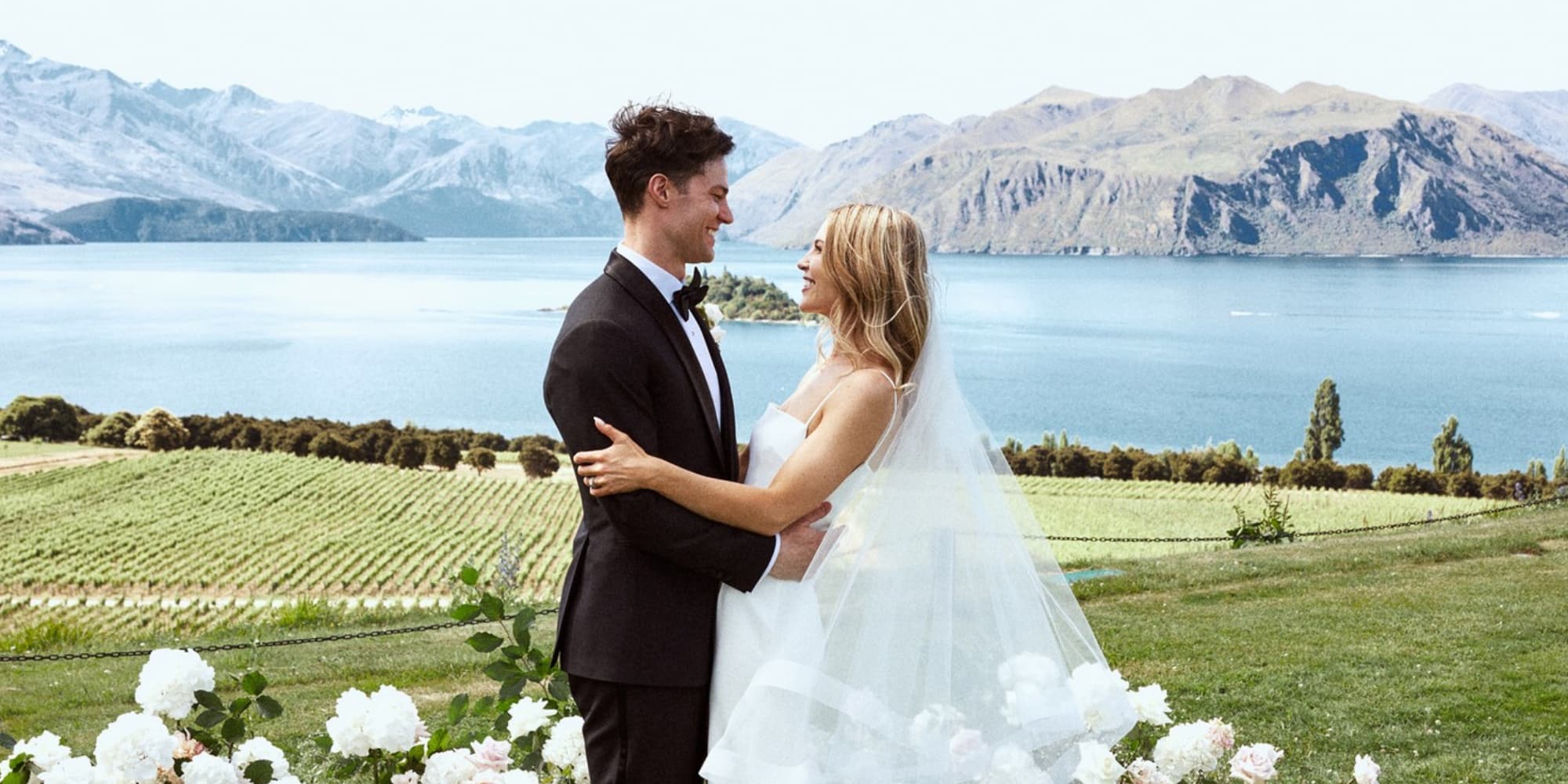 Bride and groom looking at each other with lake and mountains in background. Groom in Vitale Barberis black custom wedding tuxedo, white shirt and black bowtie. Bride in sleeveless wedding dress.