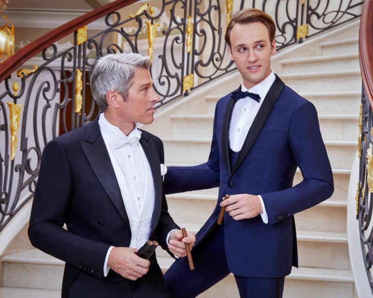 Two models in tuxedos. One in a full tail black morning suit with white pique vest. One in royal blue solid with black lapel and matching U shaped vest.