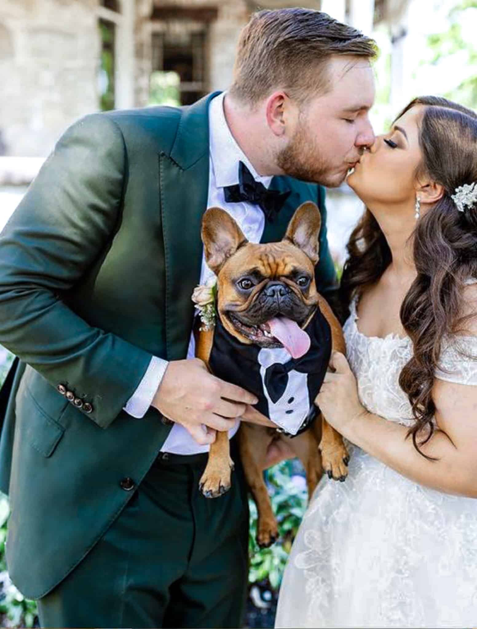 Bride and groom kissing while holding dog. Groom wearing green bespoke wedding suit. Bride in wedding dress.