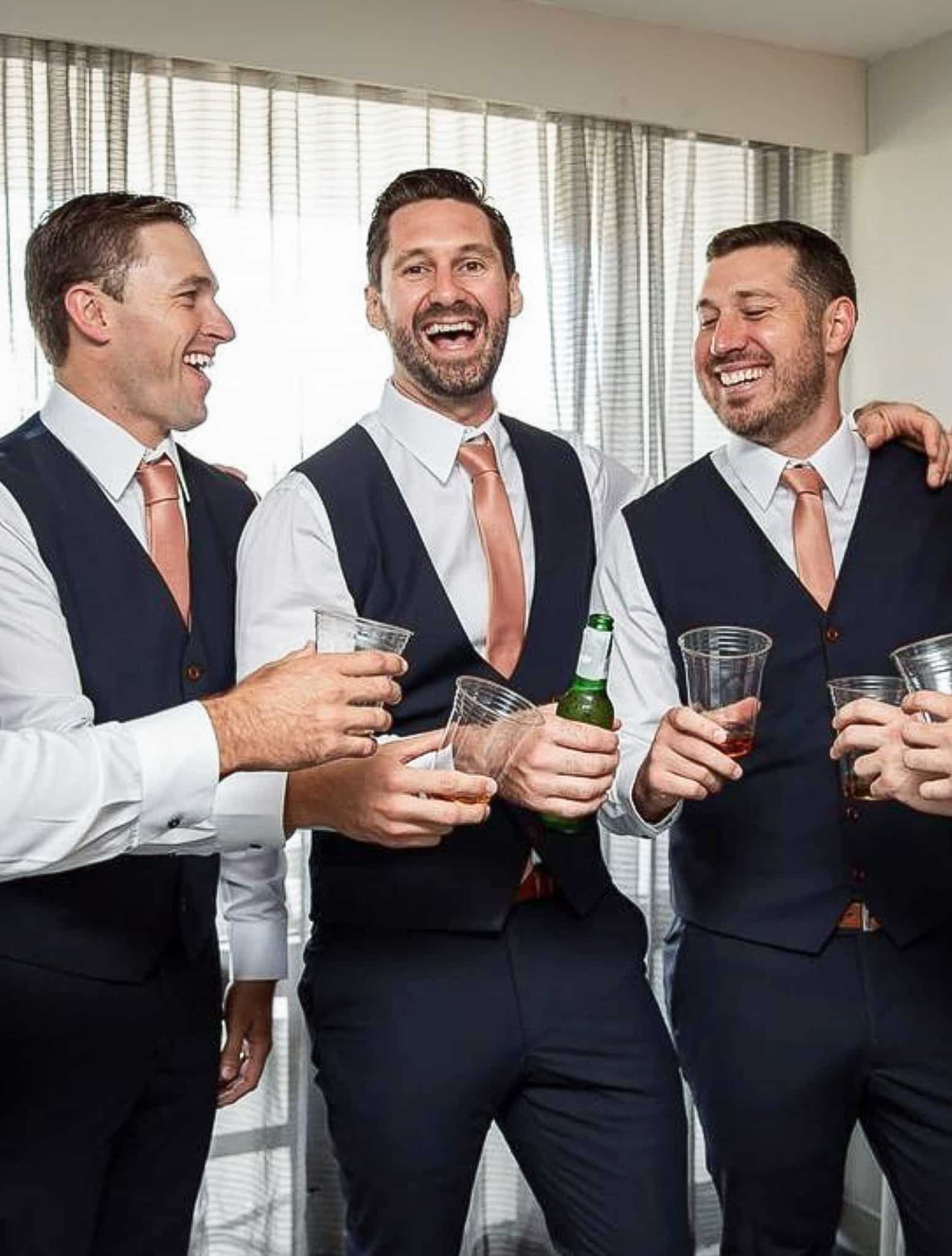 Three groomsmen holding toast glasses. All 3 wearing bespoke vests with white shirts and solid orange ties.