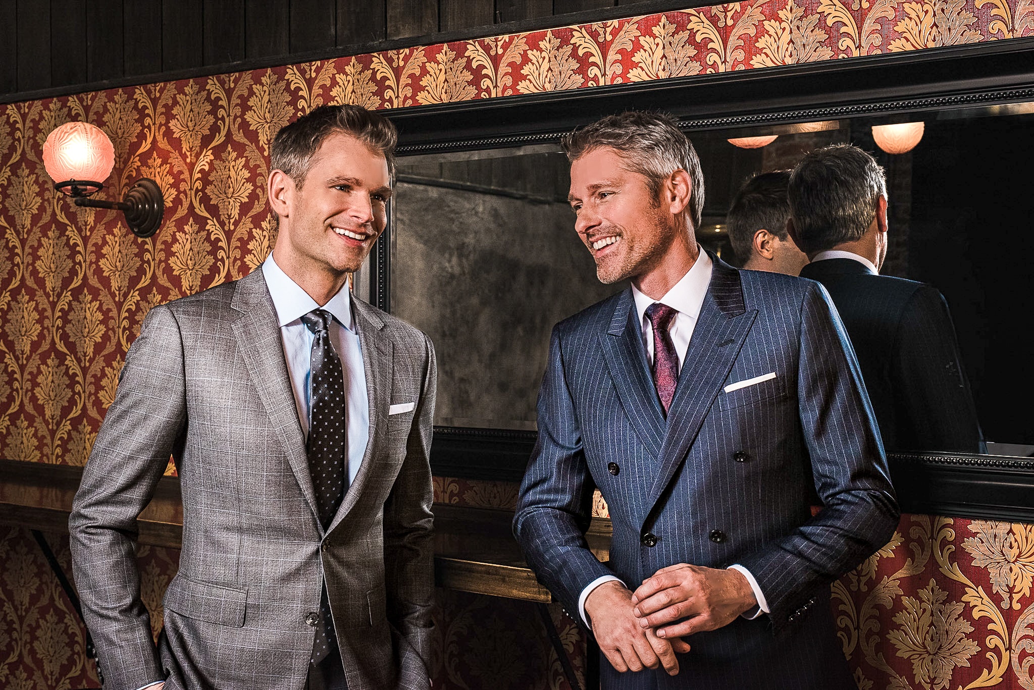 Two models wearing suits in hotel lobby. One wearing grey overplaid custom suit with blue shirt and grey polka dot tie. One wearing blue pinstripe custom suit with white shirt and red paisley tie.