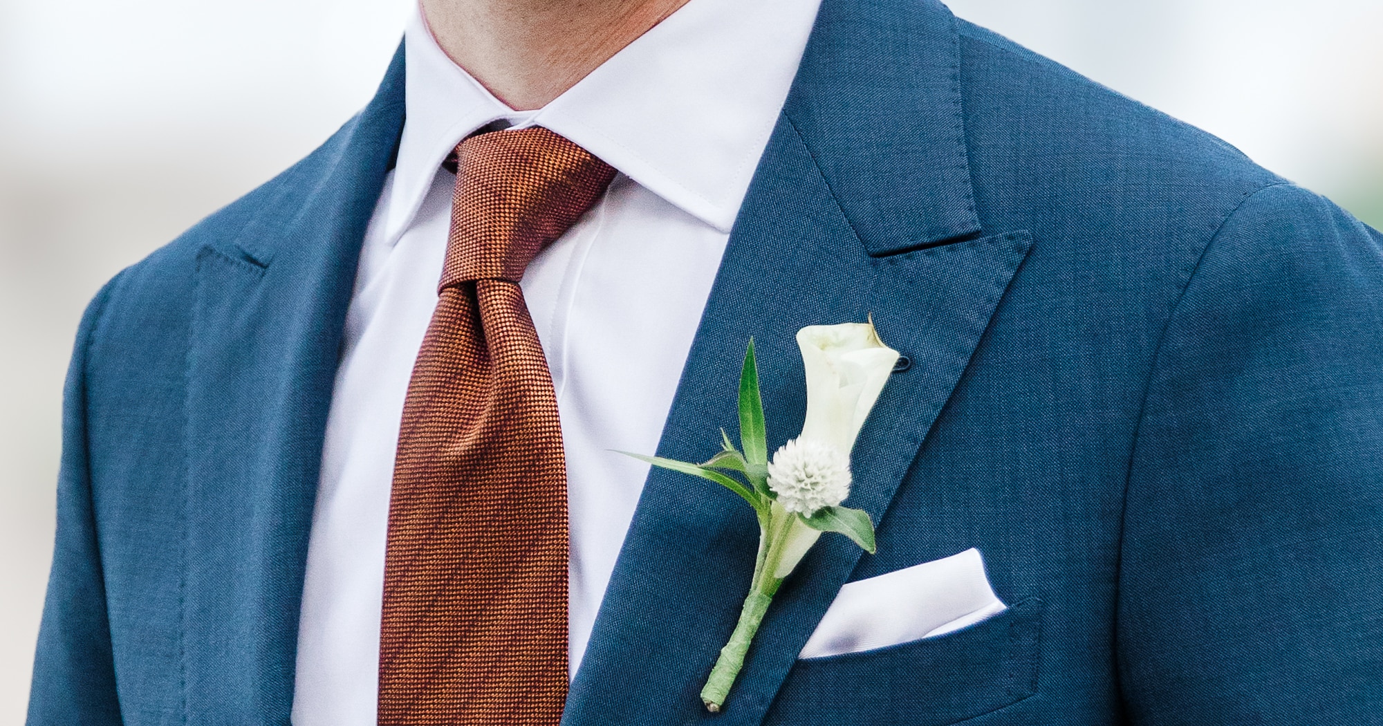 Close up of groom in medium blue solid bespoke wedding suit with peak lapel and pick stitching. White shirt and orange tie.