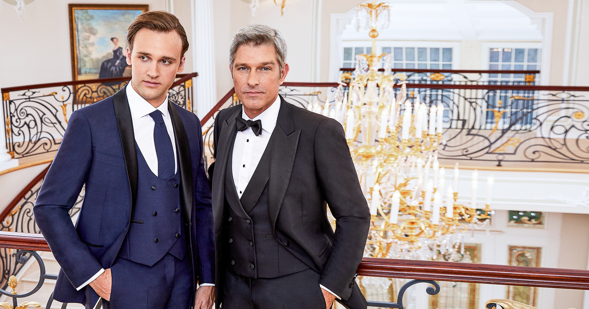 2 models wearing bespoke wedding tuxedos. One wearing black single breasted peak lapel and one wearing 3 piece blue with U shaped vest. Standing at bottom of winding staircase smoking cigars.