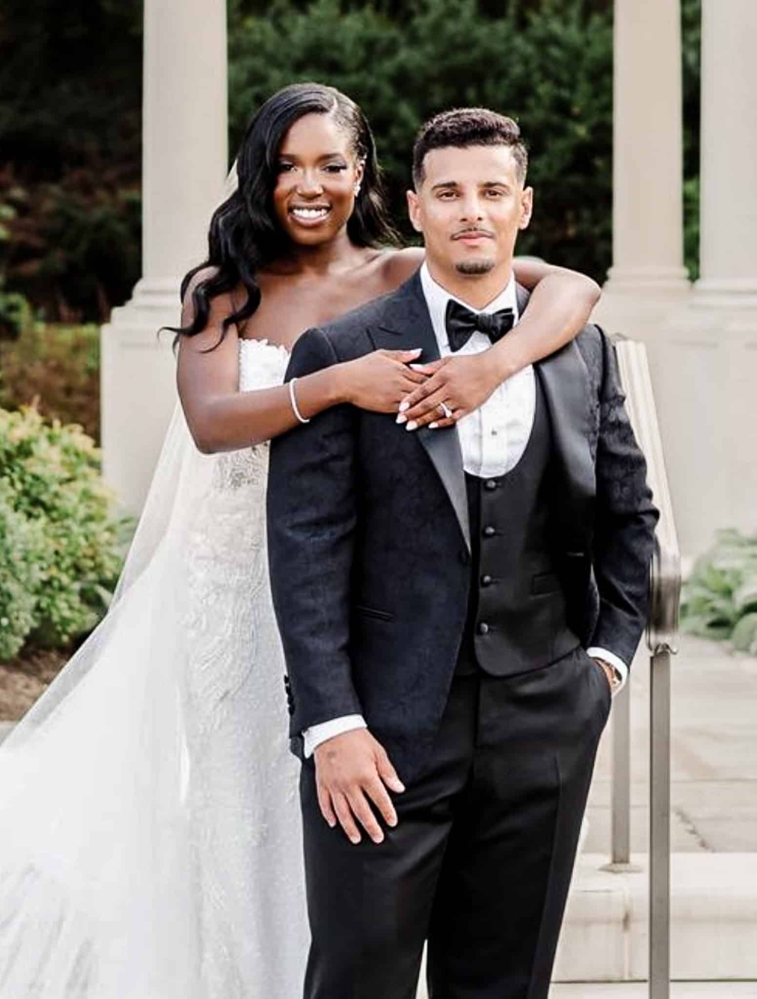 Bride with arms around groom. Groom wearing Reda black three piece tuxedo with 5 button u shaped vest, white shirt and black bowtie.