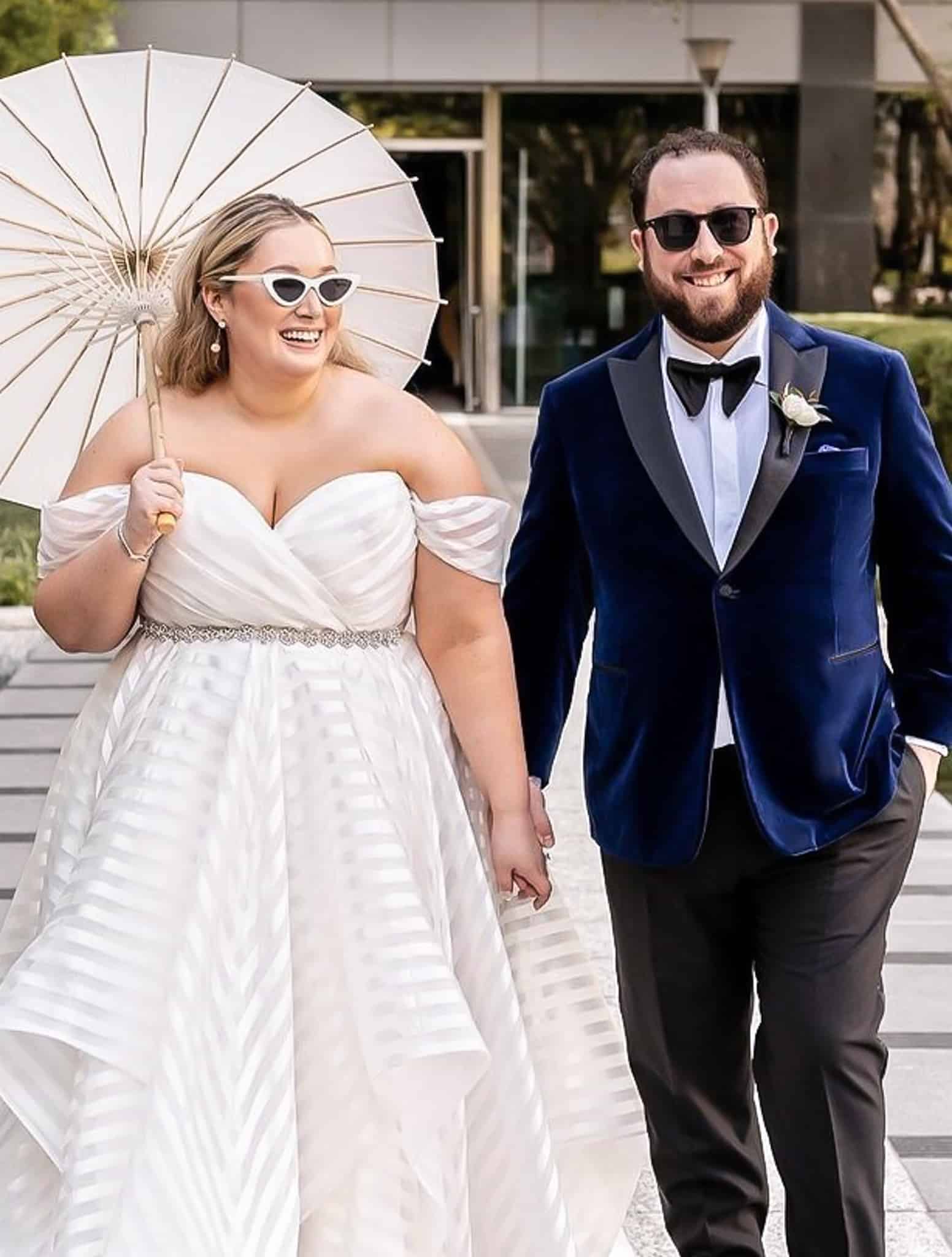 Bride & groom. Groom navy velvet dinner jacket with black satin peak lapel and charcoal trousers. White shirt and black bowtie. Bride outdoors holding umbrella.