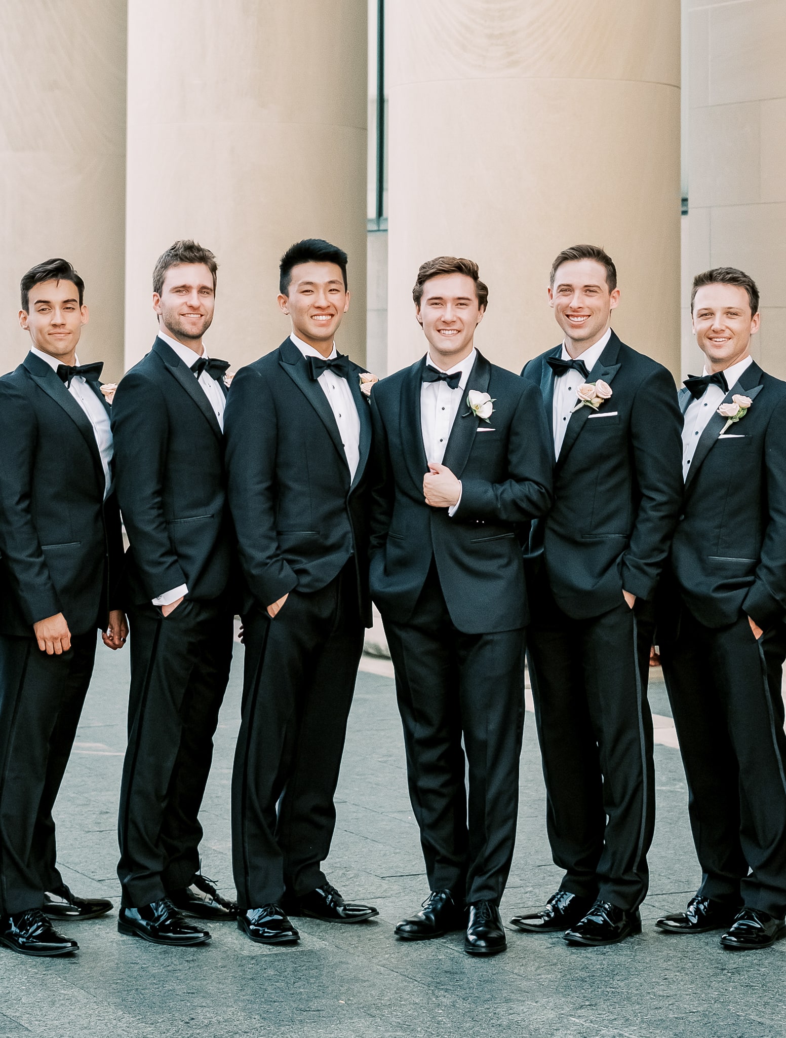 Eight groomsmen standing together wearing classic black custom tuxedos all with white shirts and black bowties.