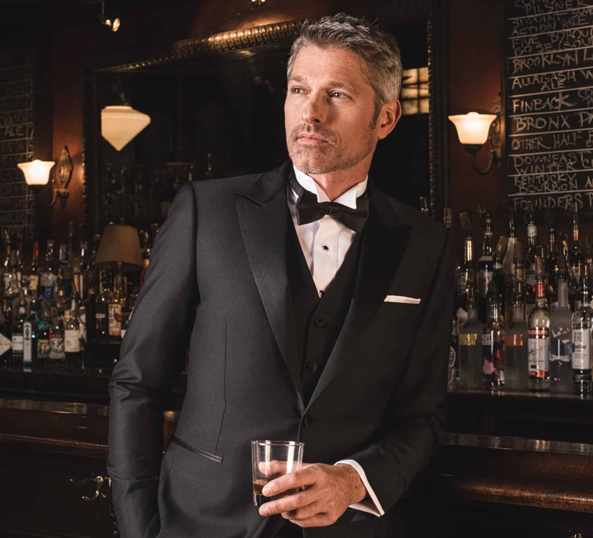 Model at bar in vested custom wedding tuxedo in black with white shirt and black bowtie. Fabric by Zegna.