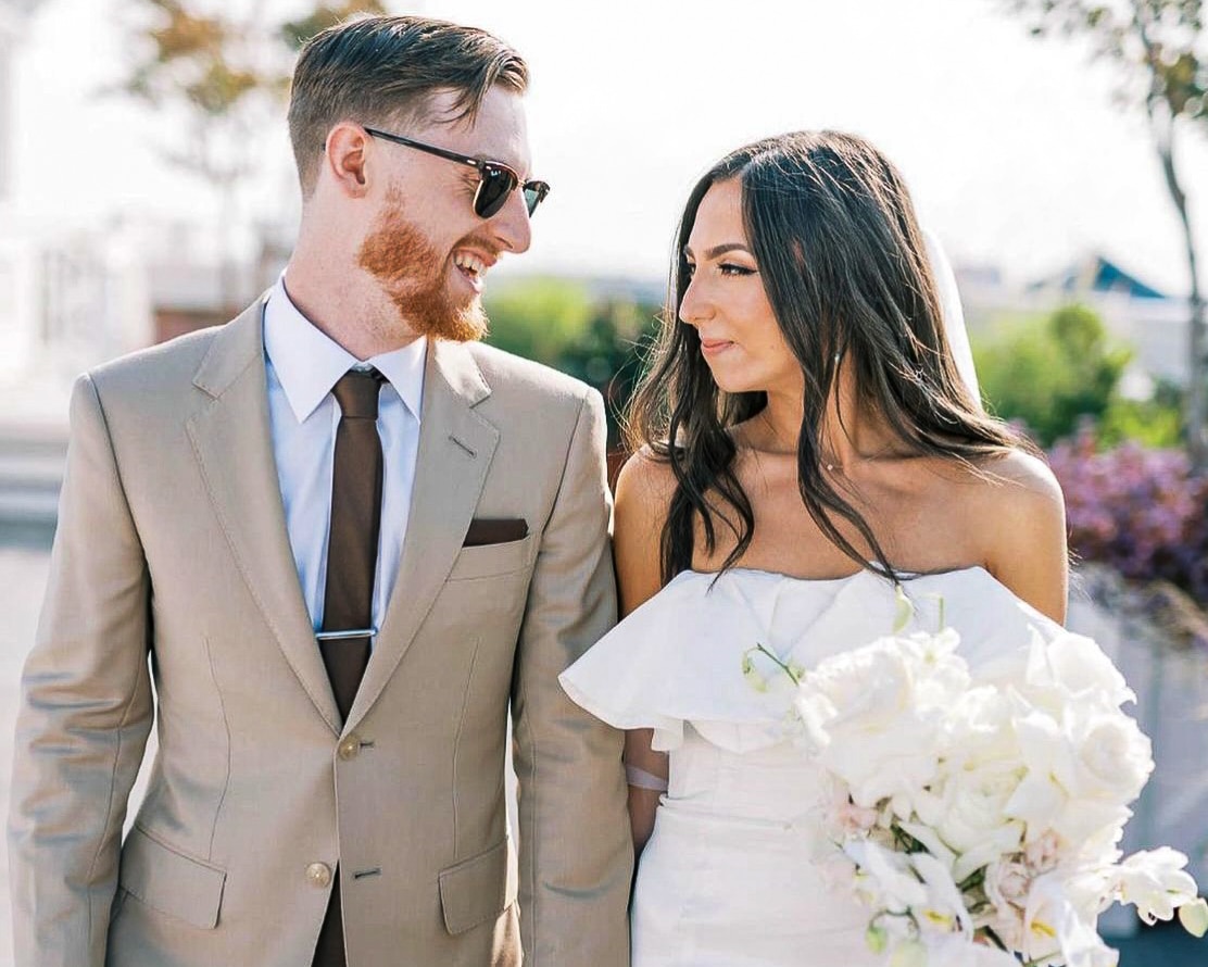 Groom and bride-groom wearing tan suit with white shirt and dark tan tie. Bride in low cut wedding dress holding flowers.