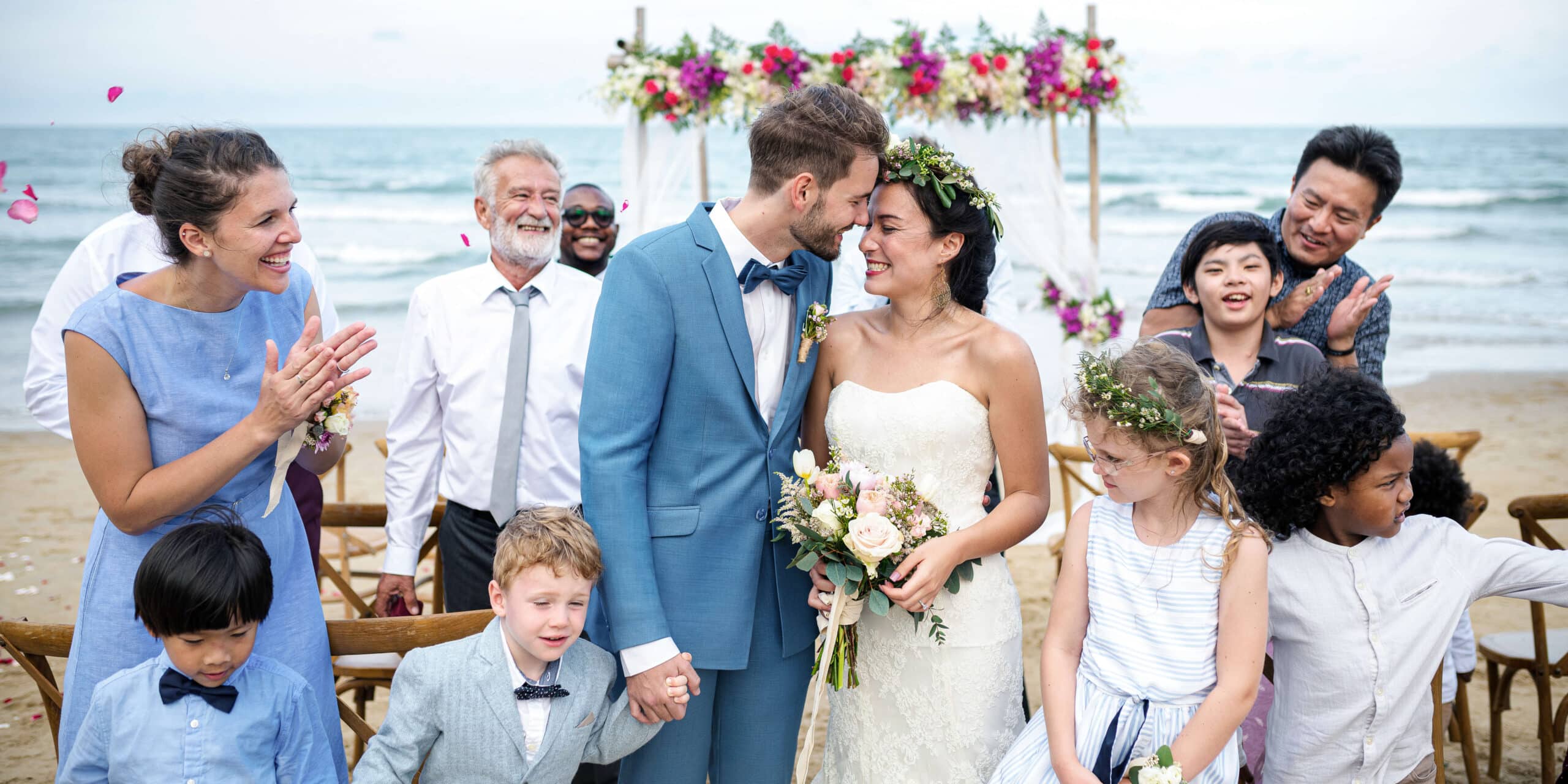 Groom getting ready to kiss bride on beach with ocean in background. Groom wearing light blue Zegna custom wedding suit with light blue bowtie. Bride in low cut wedding dress.