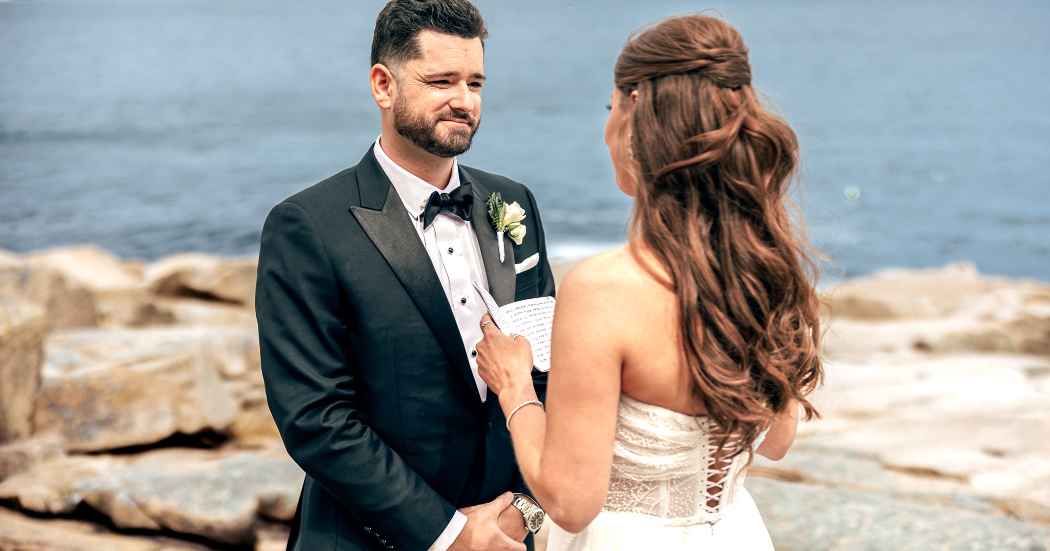 Bride reading vows to groom near rocks in front of ocean. Groom in Vitale Barberis custom tailored wedding tuxedo. Bride in off shoulder wedding dress.