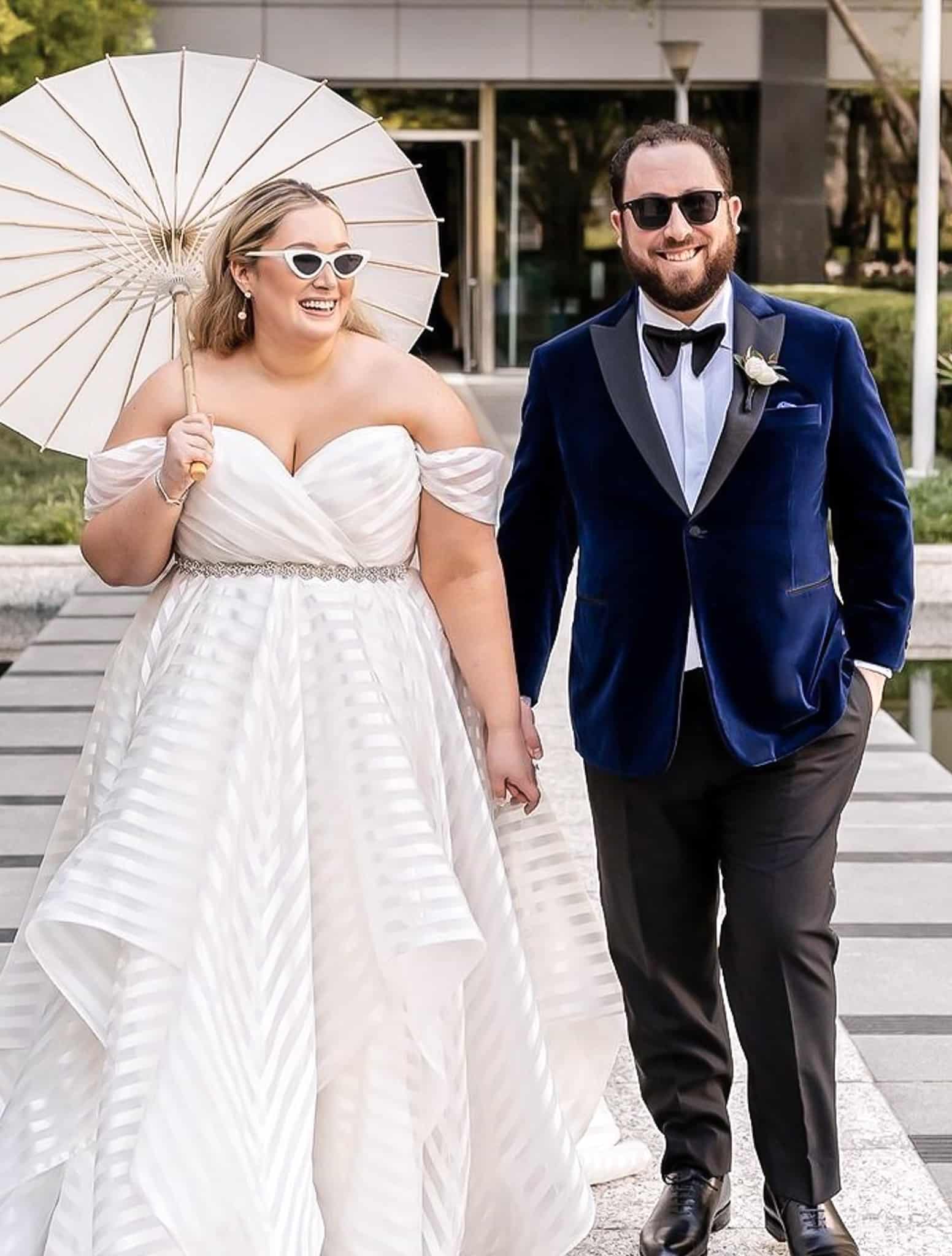 Bride and groom holding hands outdoors. Groom in Drago navy custom tailored tuxedo jacket with black peak lapel, black custom trousers, white shirt and navy bowtie. Bride holding umbrella.