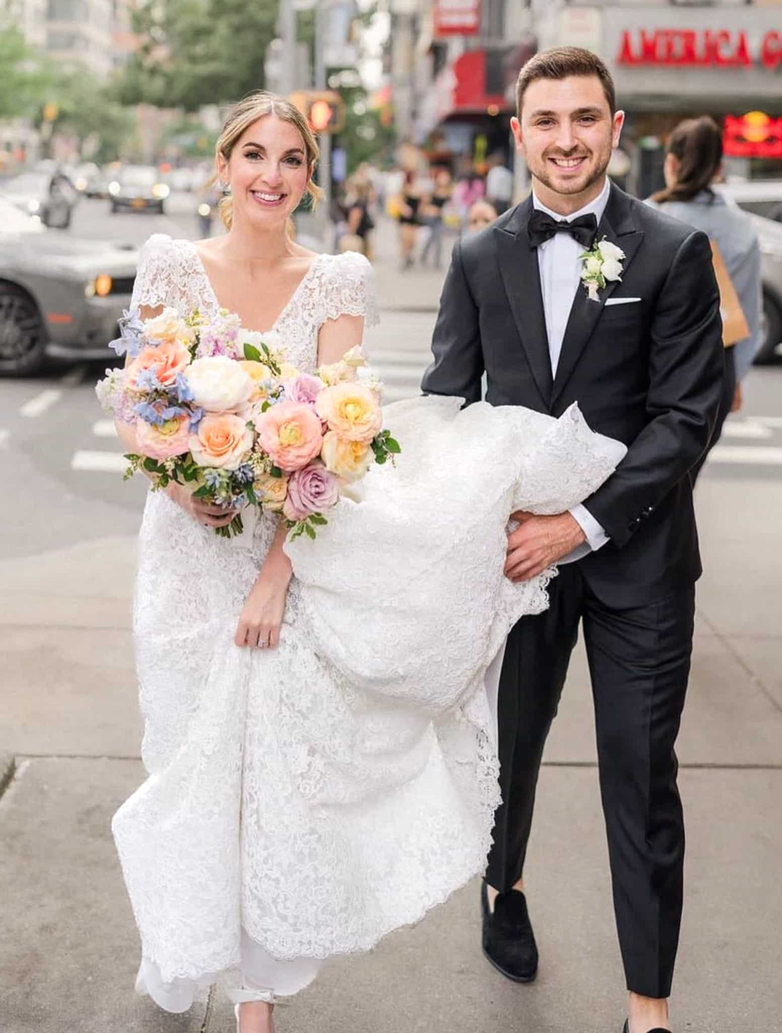 Groom holding up brides wedding dress from bottom wearing Loro Piana black classic wedding tuxedo with white shirt and black bowtie.