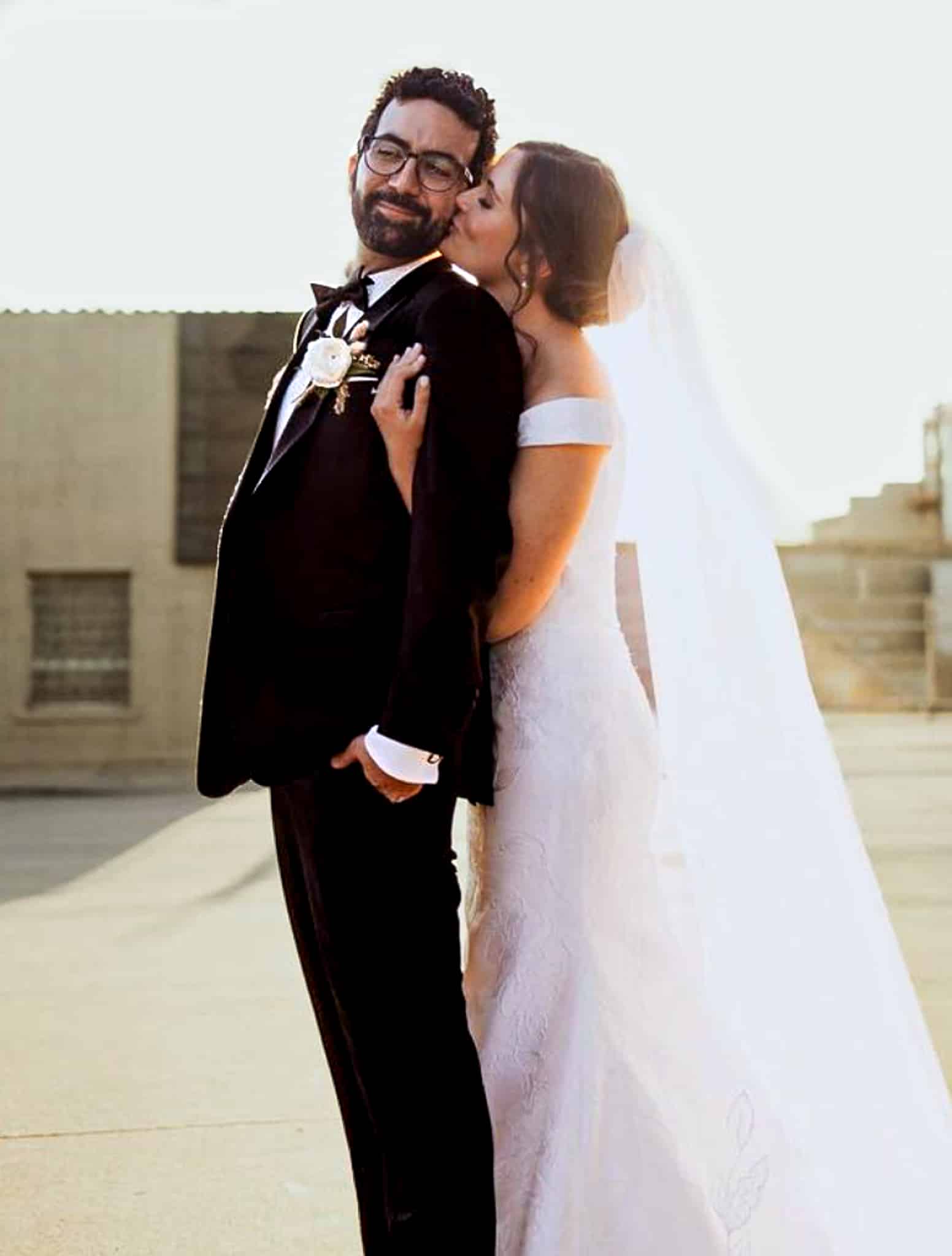Bride embracing groom from behind with kiss on cheek. Groom wearing bespoke black classic tuxedo with white shirt and black bowtie. Bride in classic wedding dress.