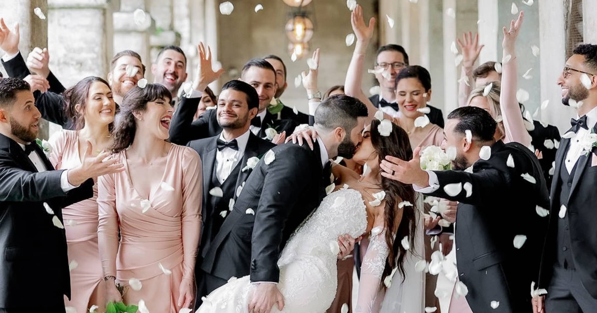 Groom kissing bride in middle of their wedding party guests. Groom wearing bespoke wedding tuxedo, bride in classic wedding dress.