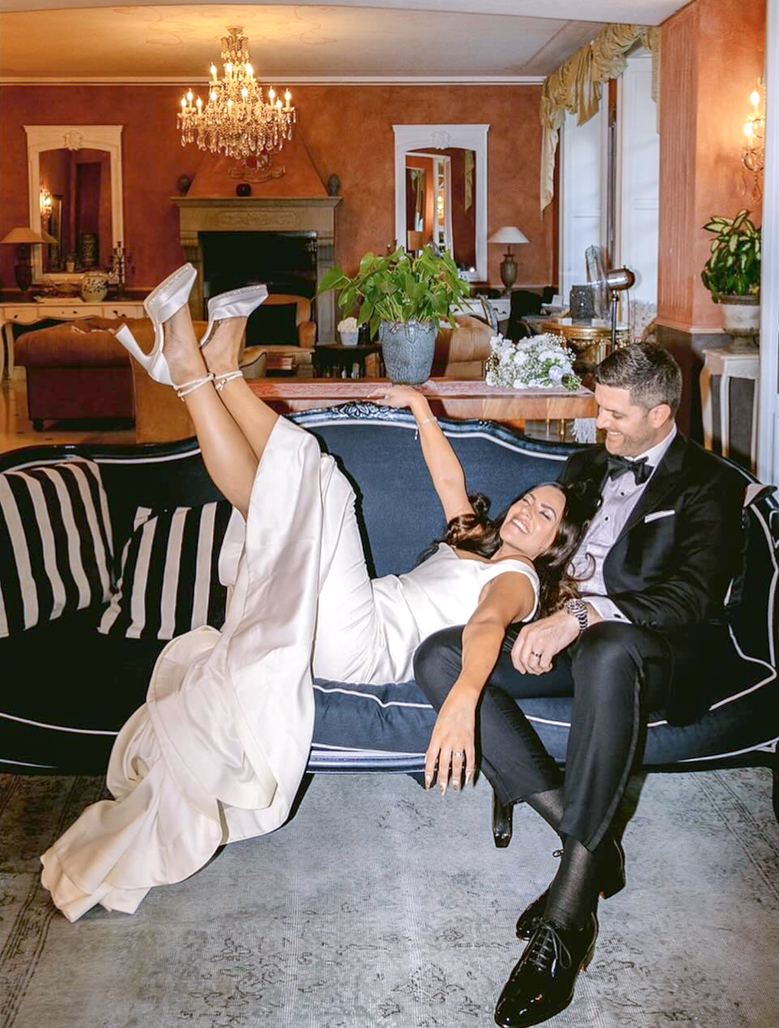 Groom sitting on couch with bride lying on his lap and legs up in air, wedding dress covering knees. Groom in classic custom wedding tuxedo.
