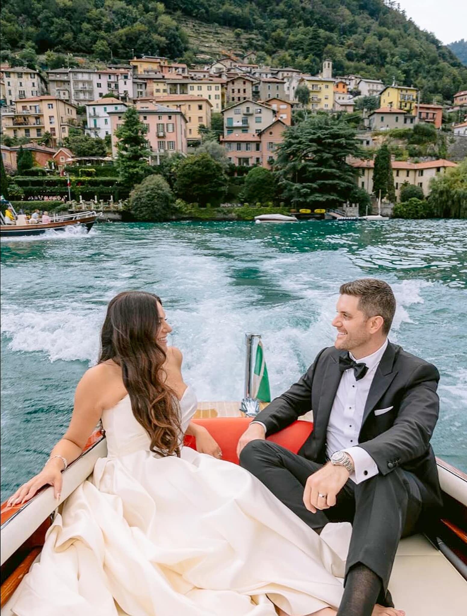 Bride and groom in boat sailing away with Italian beach houses in background. Groom wearing black peak lapel bespoke wedding tuxedo with black satin, white shirt and black bowtie. Bride in sleeveless wedding dress.