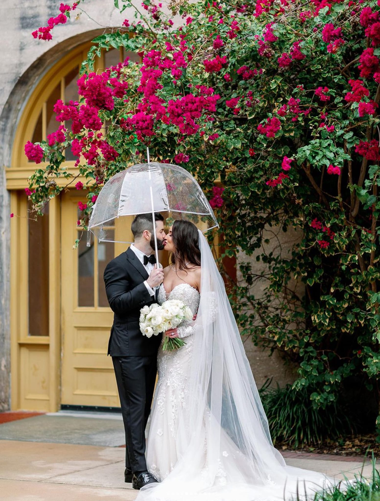 Bride and groom kissing under umbrella outside famous Greek restaurant. Groom wearing black custom wedding tuxedo with white shirt and black bowtie. Bride in classic wedding dress.