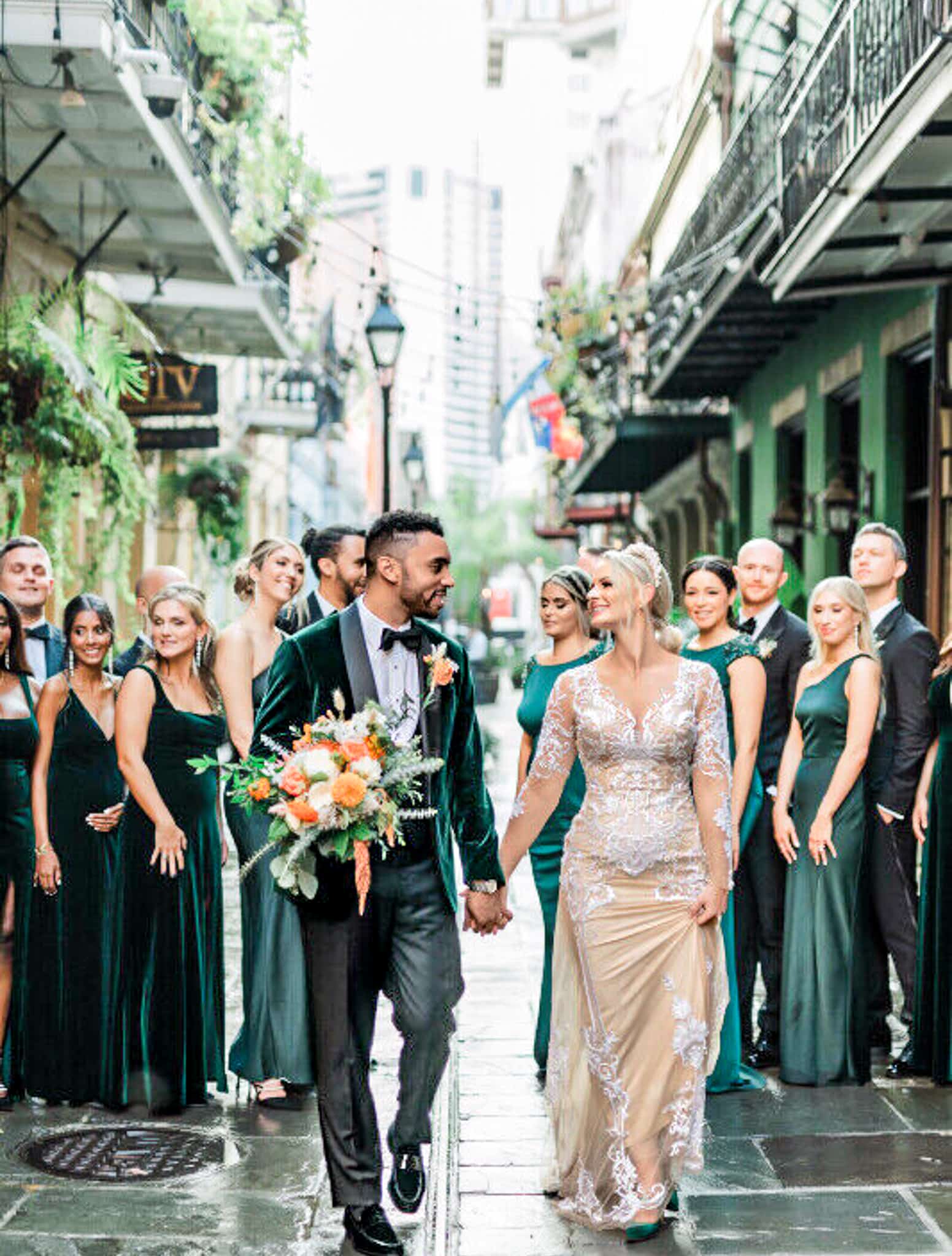 Bride and groom walking hand in hand with wedding party in background. Groom wearing Holland & Sherry green velvet vested bespoke tuxedo with black satin shawl lapel, white shirt and black bowtie. Bride wearing elegant off white wedding gown.
