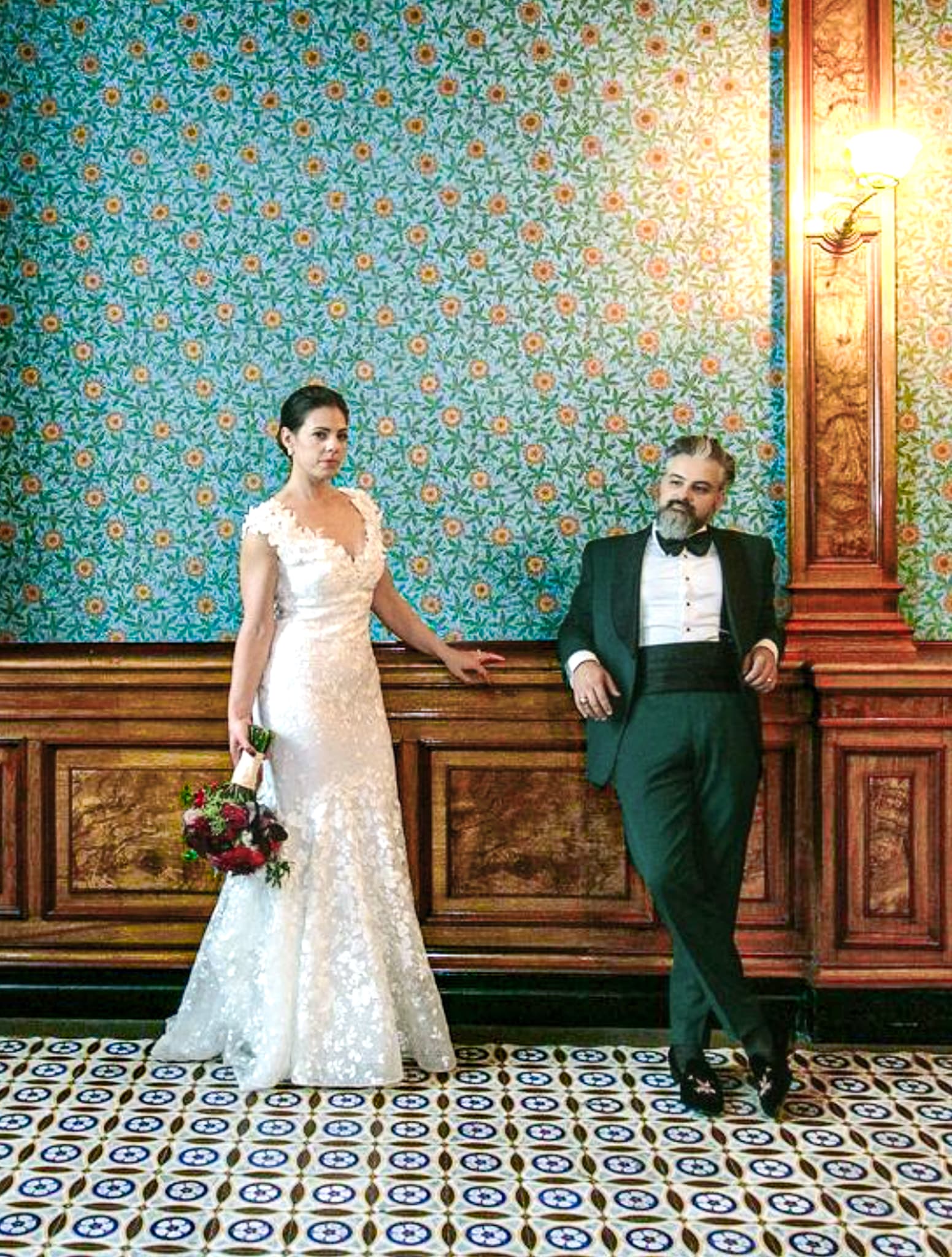 Bride and groom leaning against wall. Groom wearing green two piece wedding tuxedo from Loro Piana fabric, white shirt and black bowtie and black cummerbund. Bride in very simple sleeveless wedding dress.