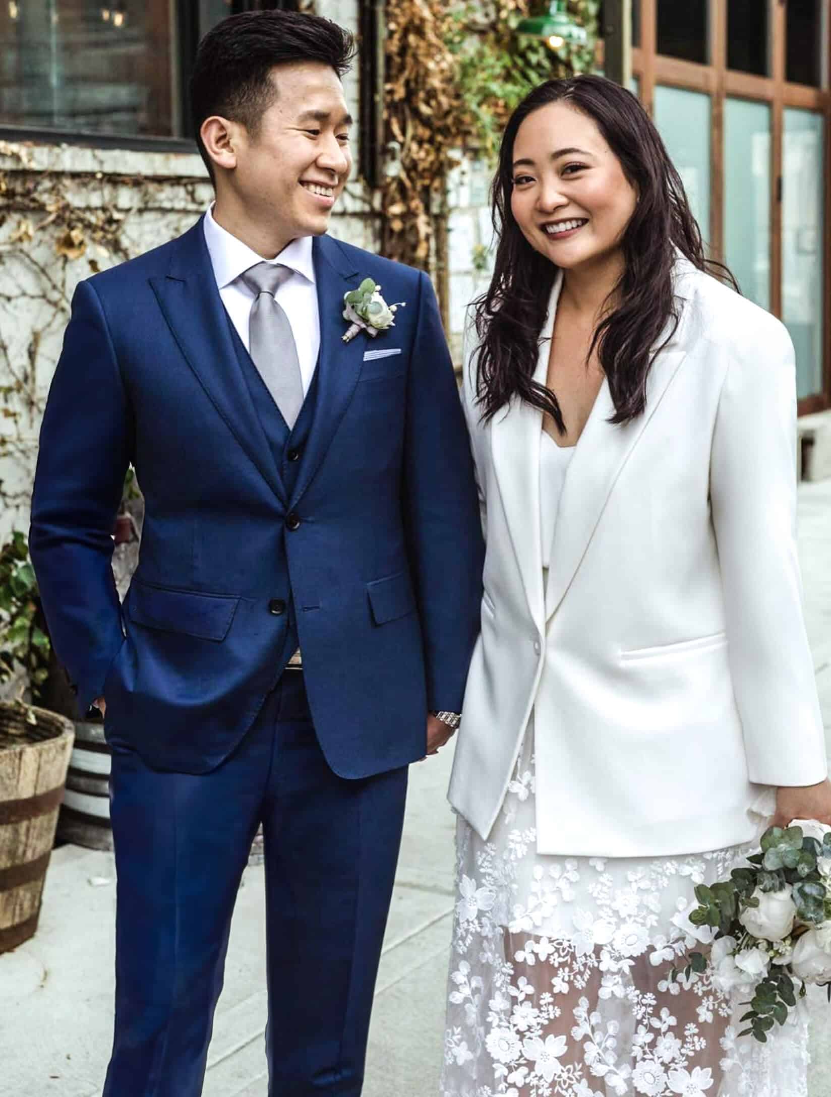 Bride and groom outdoors. Groom in navy 3 piece bespoke wedding suit with white shirt and light grey long tie, Bride in simple blazer and long skirt.