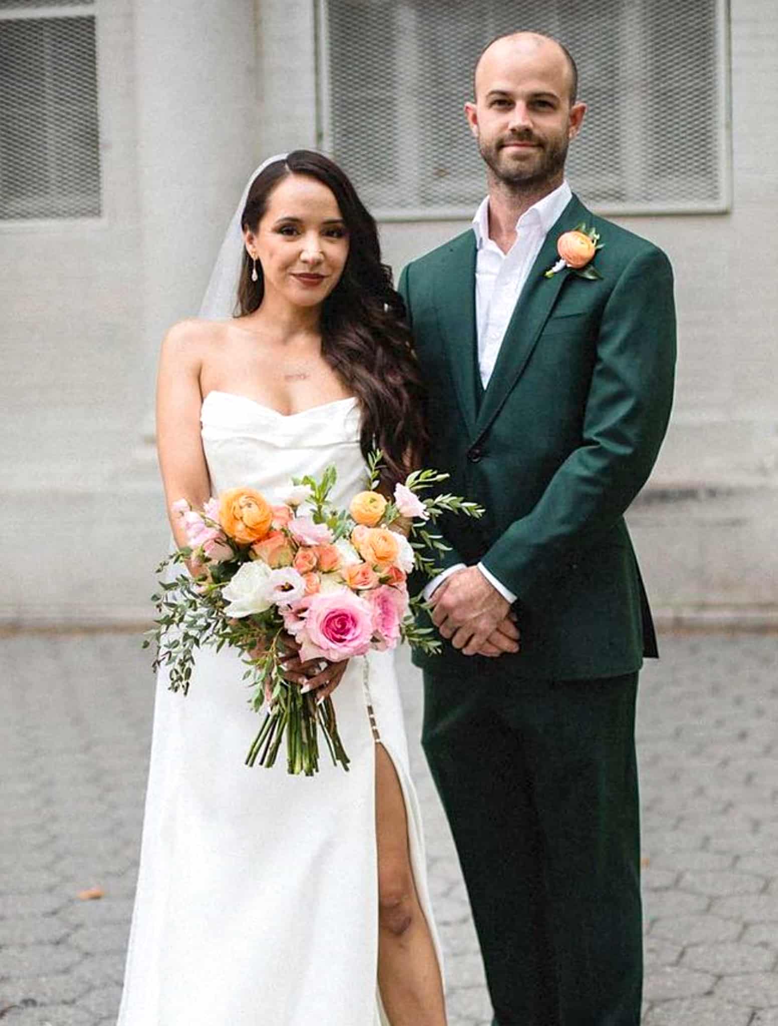 Bride and groom side by side. Groom wearing hunter green custom wedding suit with open collar white shirt. Fabric for suit by Zegna. Bride in low cut with high side slit wedding dress.