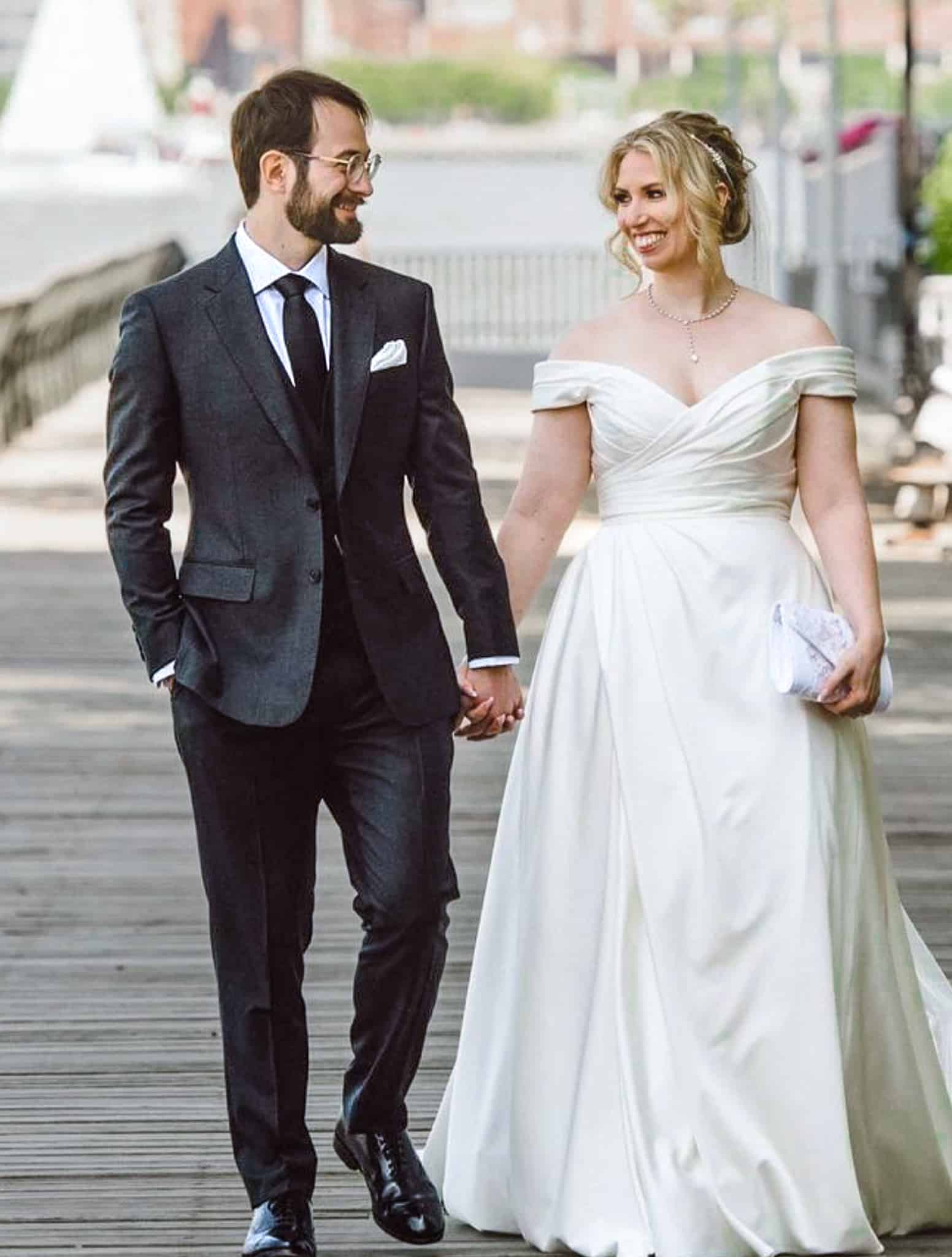 Bride and groom outdoors holding hands. Groom wearing dark charcoal custom suit with white shirt and black tie. Bride in full wedding dress.