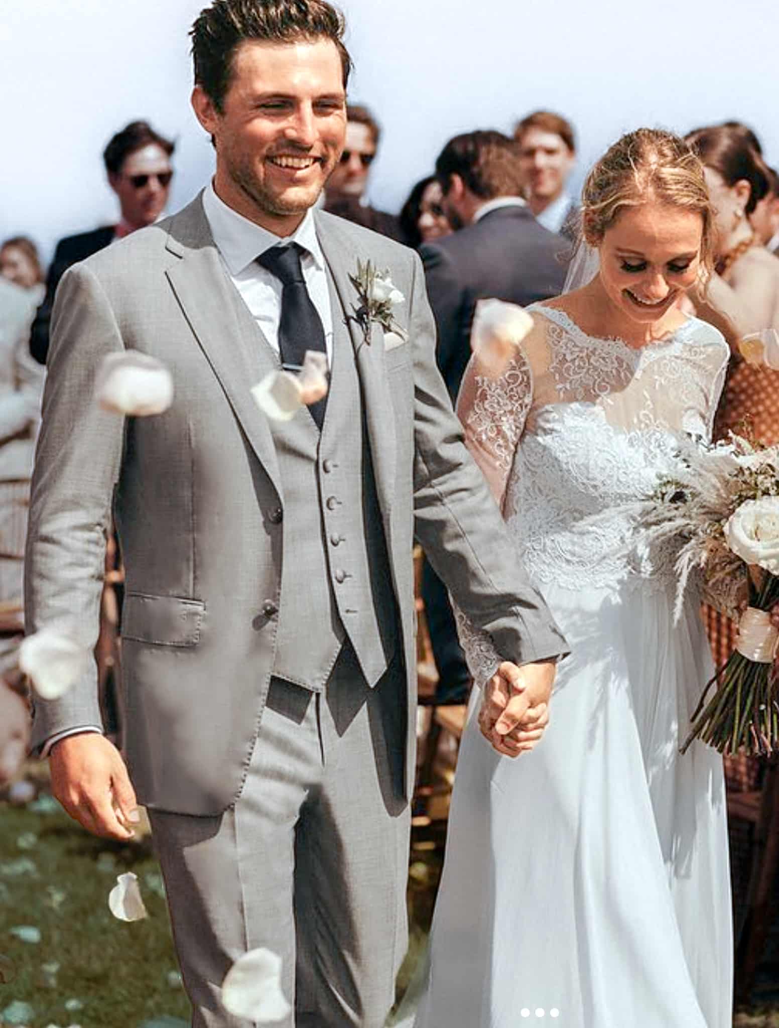 Bride and groom holding hands walking down aisle. Groom in light grey three piece bespoke wedding suit with matching vest with shawl lapel, white shirt and long tie charcoal color. Bride in classic wedding dress.
