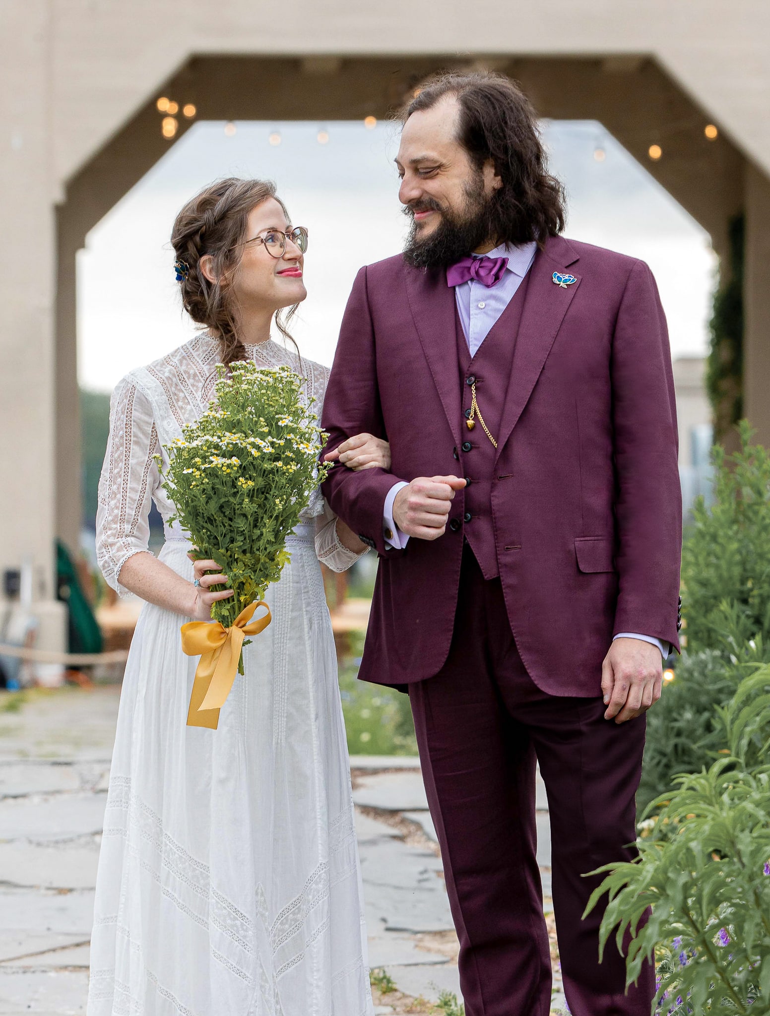 Bride & groom arm in arm outdoors. Groom wearing wine color bespoke wedding suit with matching wine vest, white shirt and black bowtie. Bride wearing high cut simple wedding dress.