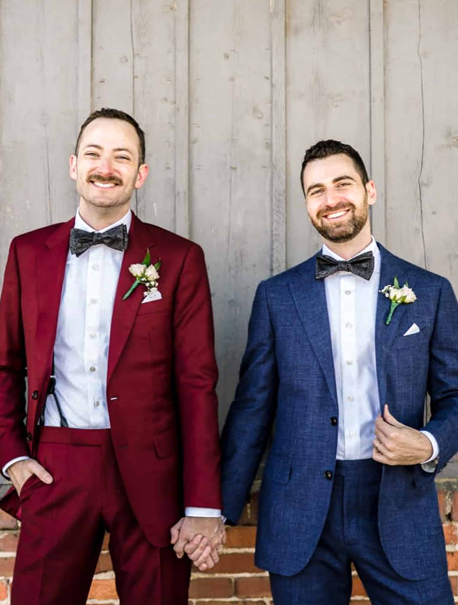 Groom and groom holding hands. One wearing solid dark blue custom wedding suit with white shirt and wide blue bowtie. Other wearing red solid bespoke wedding suit with white shirt and oversized black bowtie.
