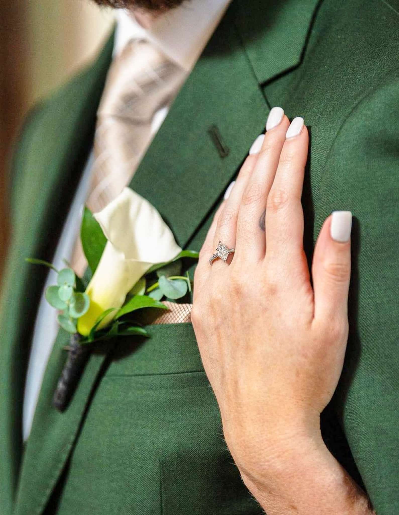 Extreme closeup of brides hand on grooms front chest near breast pocket. Groom wearing green custom wedding suit with white shirt and off white satin long tie.