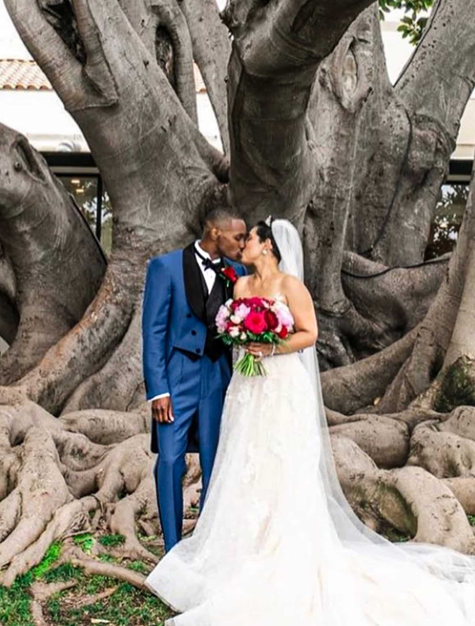 Bride and groom kissing under oak tree. Groom in blue full dress bespoke tails with black shawl satin lapel. Bride in classic wedding dress.