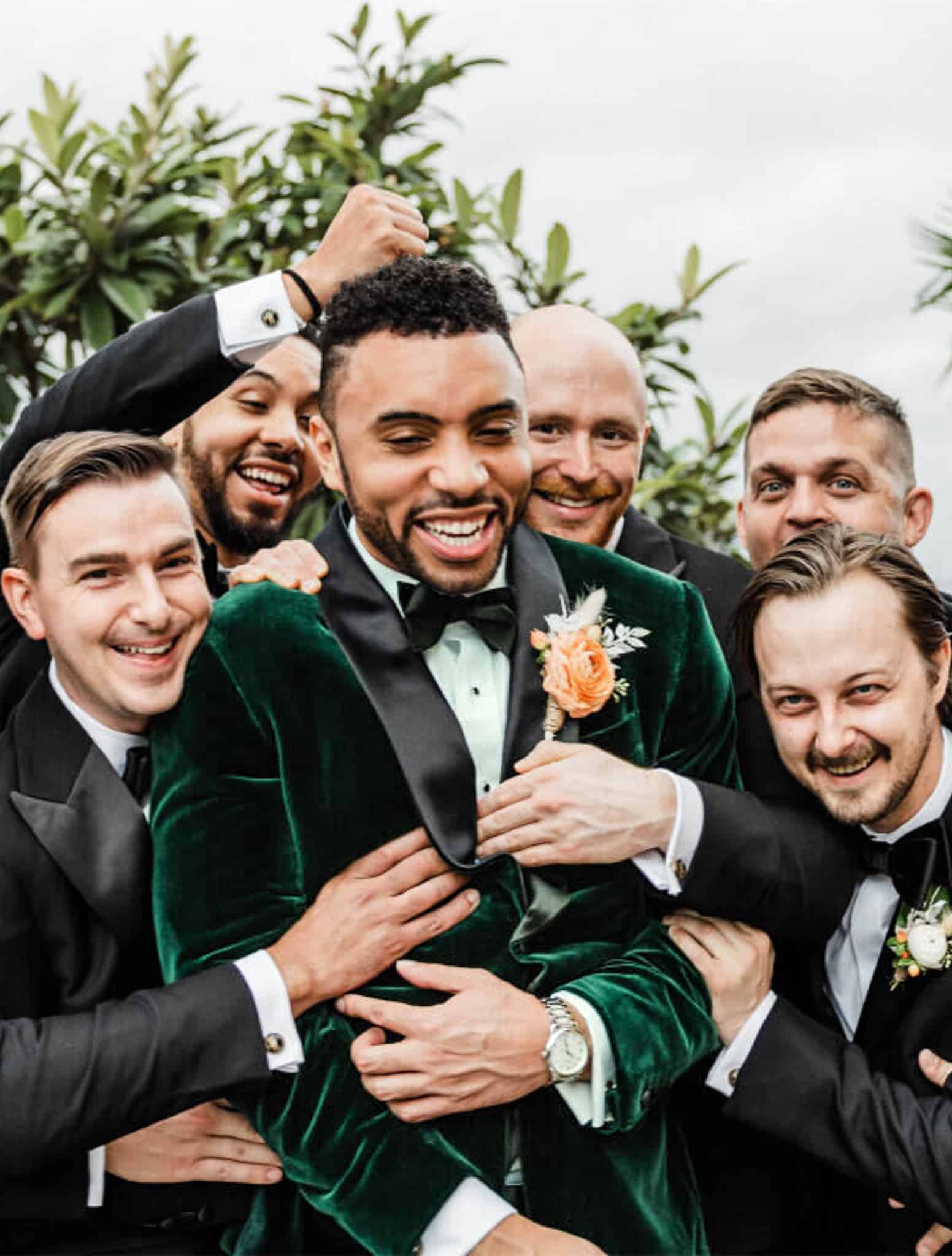 Five groomsmen in classic black tuxedo grabbing and hugging groom. Groom wearing Marzoni green velvet wedding tuxedo with black shawl lapel.
