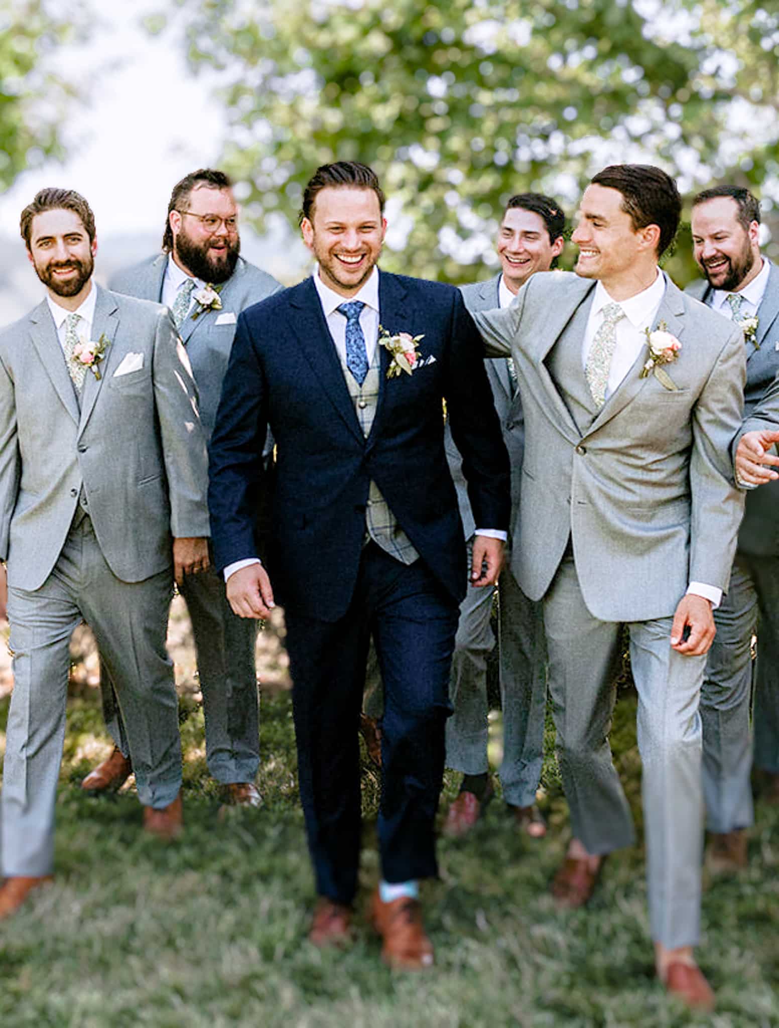 Five groomsmen in light grey custom wedding suits walking behind groom who is wearing navy blue custom tailored wedding suit with light grey and blue windowpane vest.