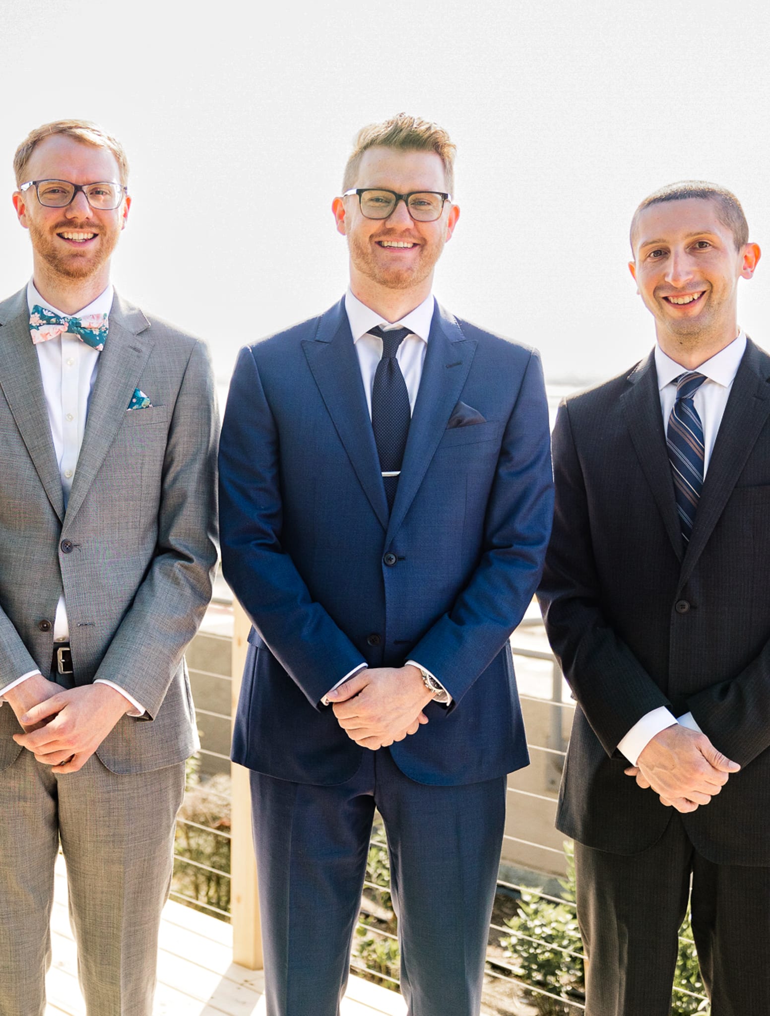 Three groomsmen all wearing wedding suits standing side by side outdoors.