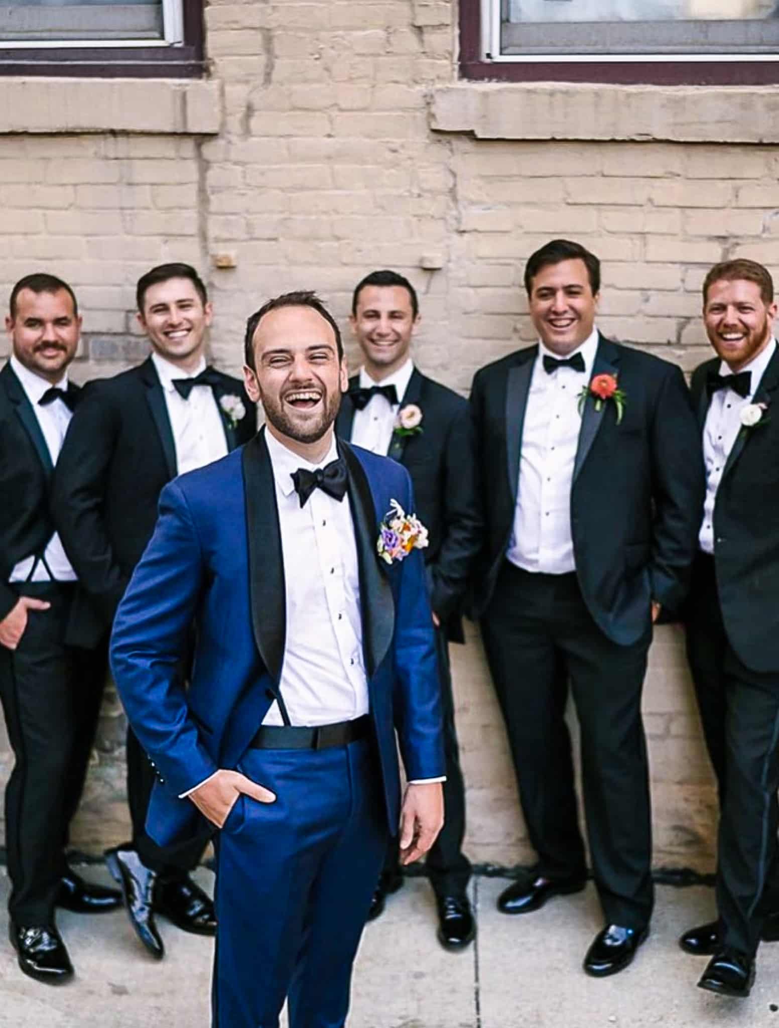 Five groomsmen standing behind groom in black custom tailored tuxedos. Groom wearing blue solid sharkskin tuxedo with black shawl lapel.