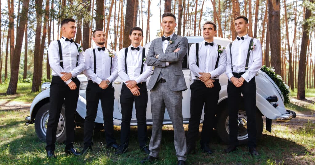 5 groomsmen with white shirts and black bowties without jackets on standing behind groom who is wearing a grey tweed custom wedding suit with a white tuxedo shirt and black bowtie.