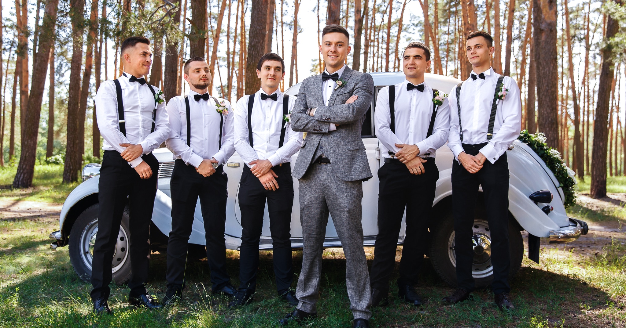 5 groomsmen with white shirts and black bowties without jackets on standing behind groom who is wearing a grey tweed custom wedding suit with a white tuxedo shirt and black bowtie.